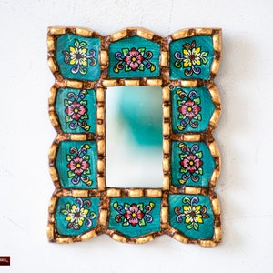 May include: A small, decorative mirror with a teal frame. The frame is intricately carved and painted with gold accents and colorful floral designs.
