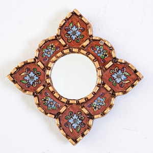 May include: A decorative, handcrafted mirror with a round central mirror and a unique, ornate frame. The frame is a warm brown color with gold accents and features a floral design with blue, yellow, and green flowers. The mirror is designed to be hung on a wall.