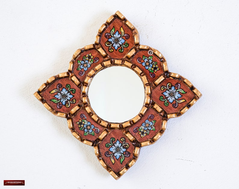 May include: A decorative, handcrafted mirror with a round central mirror and a unique, ornate frame. The frame is a warm brown color with gold accents and features a floral design with blue, yellow, and green flowers. The mirror is designed to be hung on a wall.