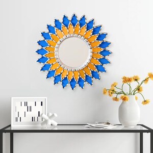 K&ouml;nnte beinhalten: Ein runder Wandspiegel mit silbernem Rahmen und einem dekorativen Sonnenstrahl-Design. Der Sonnenstrahl besteht aus blauen und gelben Glasteilen. Der Spiegel h&auml;ngt an einer wei&szlig;en Wand.