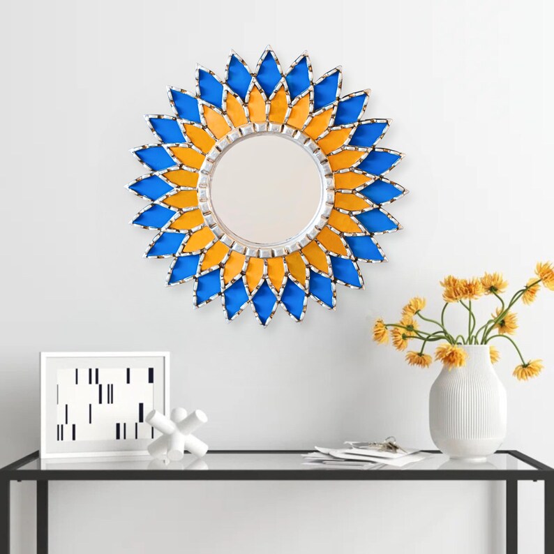 K&ouml;nnte beinhalten: Ein runder Wandspiegel mit silbernem Rahmen und einem dekorativen Sonnenstrahl-Design. Der Sonnenstrahl besteht aus blauen und gelben Glasteilen. Der Spiegel h&auml;ngt an einer wei&szlig;en Wand.