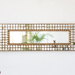 Puede incluir: Un espejo rectangular con un marco dorado hecho de pequeños cuadrados. El espejo refleja una planta con hojas verdes.