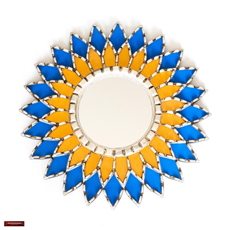 K&ouml;nnte beinhalten: Runder dekorativer Spiegel mit Sonnenblumenmuster. Der Spiegel hat einen silbernen Rahmen und ist von blauen und gelben rautenf&ouml;rmigen Glasteilen umgeben. Eine dekorative Wanddekoration.