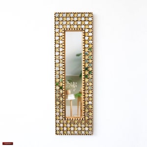 Puede incluir: Un espejo de pared rectangular con un marco de mosaico. El marco está hecho de pequeñas baldosas cuadradas en tonos dorados y plateados. El espejo refleja una habitación con una pared blanca y una pequeña mesa.