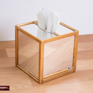 K&ouml;nnte beinhalten: Ein goldfarbener, verspiegelter Taschentuchspender mit einem wei&szlig;en Taschentuch im Inneren. Die Box ist quadratisch und hat auf allen Seiten eine verspiegelte Oberfl&auml;che. Die Box steht auf einer Holzoberfl&auml;che.