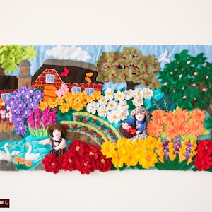 May include: A colorful felt wall hanging depicting a whimsical scene with a red barn, flowers, trees, and two figures. The figures are dressed in blue and red clothing and are holding baskets of flowers. The background is a light blue sky with white clouds.