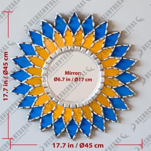 K&ouml;nnte beinhalten: Ein dekorativer Wandspiegel mit floralem Design. Der Rahmen des Spiegels ist eine Kombination aus blauen und gelben Glasteilen, die in einem Sonnenstrahlmuster angeordnet sind. Der Spiegel selbst hat einen Durchmesser von 17 cm, der Gesamtdurchmesser betr&auml;gt 45 cm.