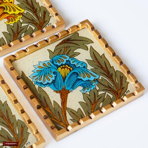 May include: Four square coasters with a gold frame and a floral design. The coasters feature a blue and yellow flower with green leaves on a cream background.