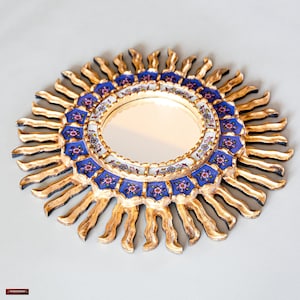 May include: A gold sunburst mirror with a blue and white floral design. The mirror is round and has a simple frame.