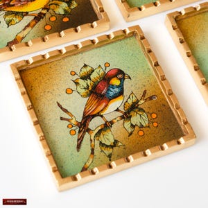 May include: Set of four square coasters with a gold frame. Each coaster features a colorful bird perched on a branch with green leaves and orange berries on a green and brown background.
