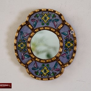 Puede incluir: Un peque&ntilde;o espejo redondo con un marco decorativo. El marco est&aacute; pintado en color morado claro con detalles dorados y dise&ntilde;os florales en amarillo, rosa y verde.