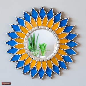 K&ouml;nnte beinhalten: Runder dekorativer Spiegel mit silbernem Rahmen und Sonnenstrahl-Design. Der Sonnenstrahl besteht aus blauen und gelben Glasteilen. Der Spiegel ist ein dekorativer Wandschmuck.
