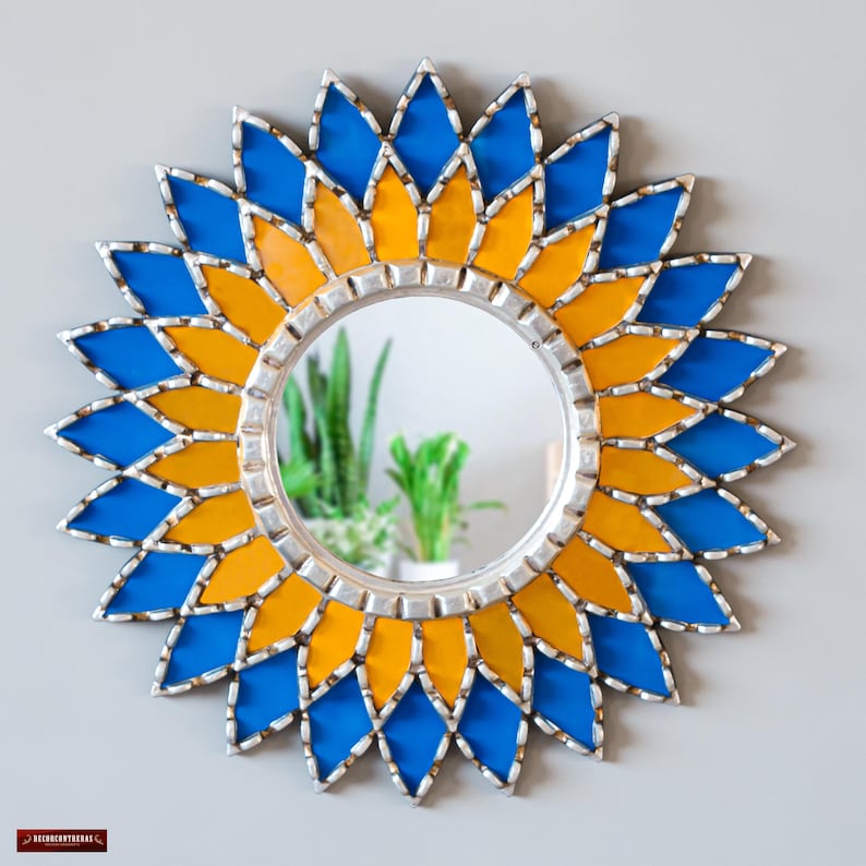 K&ouml;nnte beinhalten: Runder dekorativer Spiegel mit silbernem Rahmen und Sonnenstrahl-Design. Der Sonnenstrahl besteht aus blauen und gelben Glasteilen. Der Spiegel ist ein dekorativer Wandschmuck.