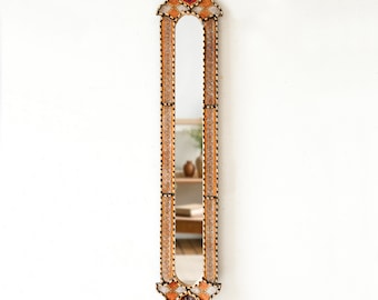 May include: A tall, narrow mirror with a decorative frame made of mosaic tiles in shades of brown, orange, red, and white. The frame features intricate geometric patterns and a pointed arch at the top.