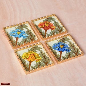May include: Four square coasters with gold trim and floral designs. Each coaster features a different colored flower with a white background. The flowers are blue, yellow, orange, and blue.