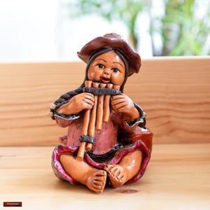 Könnte beinhalten: Eine Keramikfigur einer Person, die eine Panflöte spielt. Die Figur trägt einen braunen Hut und ein rosa-rotes Kleid. Die Figur sitzt auf einer Holzoberfläche.