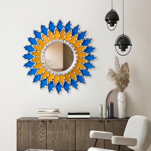 K&ouml;nnte beinhalten: Ein runder dekorativer Spiegel mit silbernem Rahmen und einem Sonnenstrahl-Design. Der Sonnenstrahl besteht aus blauen und gelben Glasteilen. Der Spiegel ist an einer wei&szlig;en Wand &uuml;ber einem Holzschrank angebracht.