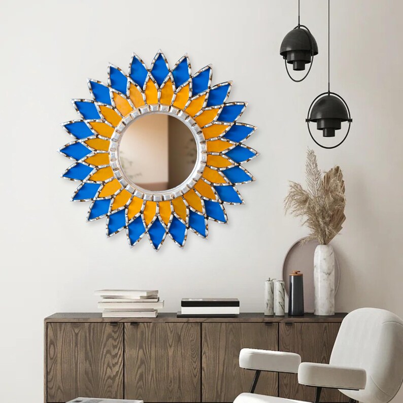 K&ouml;nnte beinhalten: Ein runder dekorativer Spiegel mit silbernem Rahmen und einem Sonnenstrahl-Design. Der Sonnenstrahl besteht aus blauen und gelben Glasteilen. Der Spiegel ist an einer wei&szlig;en Wand &uuml;ber einem Holzschrank angebracht.