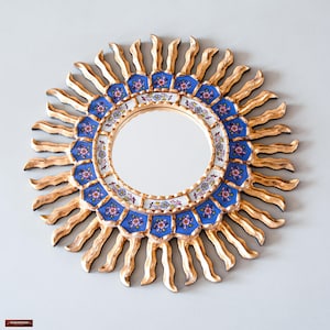 Close detail of the reverse painted glass panels of this Peruvian sunburst mirror, where pink and violet colonial flowers sit on a deep sapphire ground behind the glass, framed by hand carved wood covered in bronze leaf and softened with a nogalina aging that settles into the cuts.
