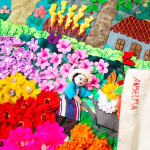May include: A detailed textile artwork depicting a garden with felt flowers in pink, yellow, red, and purple. A small figure and a house are also included. The name "ANSELMA" is embroidered in red on a white cloth, adding a personal touch to the scene.