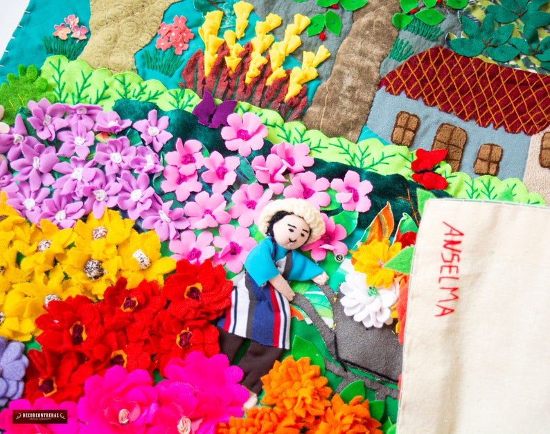 May include: A detailed textile artwork depicting a garden with felt flowers in pink, yellow, red, and purple. A small figure and a house are also included. The name "ANSELMA" is embroidered in red on a white cloth, adding a personal touch to the scene.