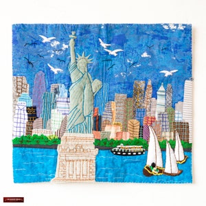 Op de afbeelding: Een kleurrijk patchwork-deken dat de skyline van New York City afbeeldt met het Vrijheidsbeeld op de voorgrond. Het deken is gemaakt van verschillende stoffen en texturen, waaronder denim, ruit en bloemenprints. De lucht is een levendige blauwe kleur met witte vogels die erboven vliegen. Het water is diepblauw met witte golven. Er zijn zeilboten in het water.