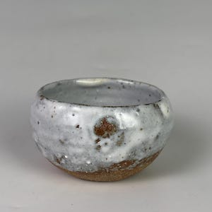 May include: A small, handmade ceramic bowl with a rounded shape. The bowl features a white glaze with brown speckles and a natural, unglazed brown base. The interior is a light gray color.