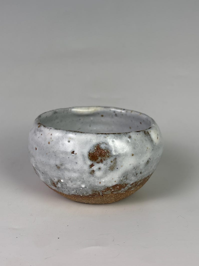 May include: A small, handmade ceramic bowl with a rounded shape. The bowl features a white glaze with brown speckles and a natural, unglazed brown base. The interior is a light gray color.