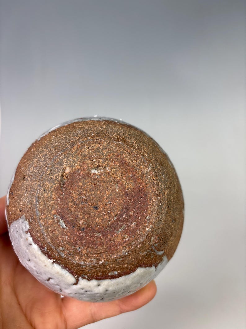 May include: A handmade ceramic bowl with a textured, brown exterior and a white glazed rim. The bowl's base features a spiral pattern, showcasing the craftsmanship and unique design of the pottery.