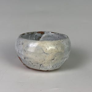May include: A small, round ceramic bowl with a speckled, off-white glaze. The bowl has a slightly irregular shape and a visible brown rim. The interior is also glazed, and the overall aesthetic is rustic and handmade.