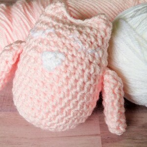 May include: A pink crocheted stuffed animal with white accents. The animal has two small arms and a round body. There are two balls of yarn in the background, one pink and one white.