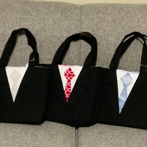 May include: Three black tote bags with handles, each designed to resemble a tuxedo. The bags feature white shirt fronts with different colored ties: gray, red, and blue. The bags are sitting on a gray couch.