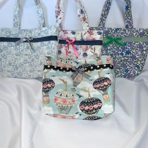 May include: Four tote bags with floral patterns and bows. The bags have different floral designs in shades of blue, pink, and purple. Each bag has a contrasting band and a bow. The bottom bag features hot air balloons.