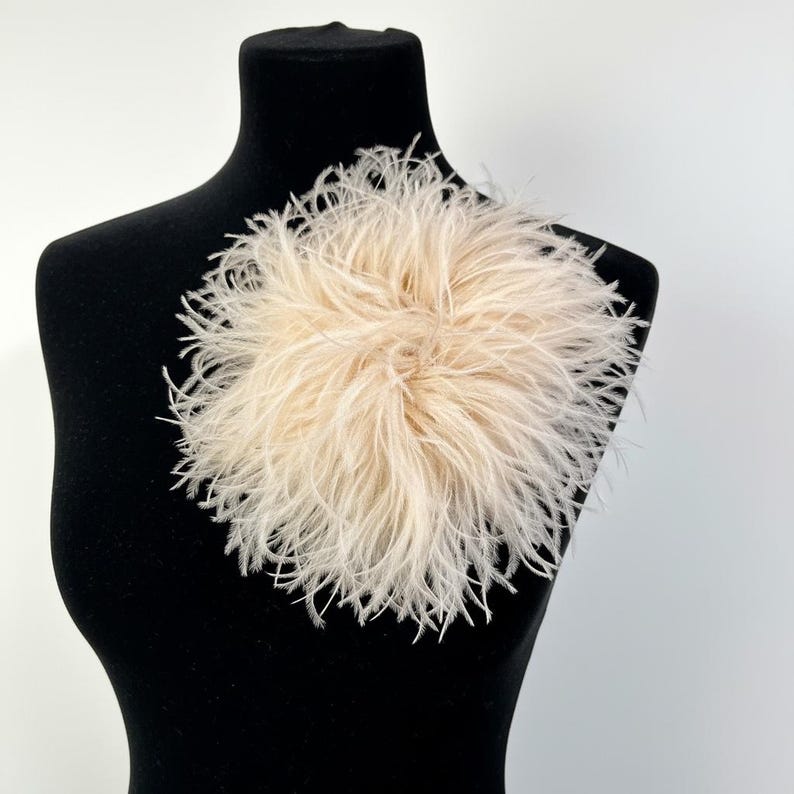 May include: A round, fluffy brooch made of light beige ostrich feathers. The delicate, wispy plumes are arranged in a dense, circular cluster, creating a soft, voluminous texture that radiates outward from the center of the accessory.