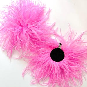 May include: Two bright pink feather accessories, each featuring a circular black base with a silver metal clip attached to the center. The soft, fluffy plumes radiate outward from the dark circular centers, creating a voluminous, textured appearance.