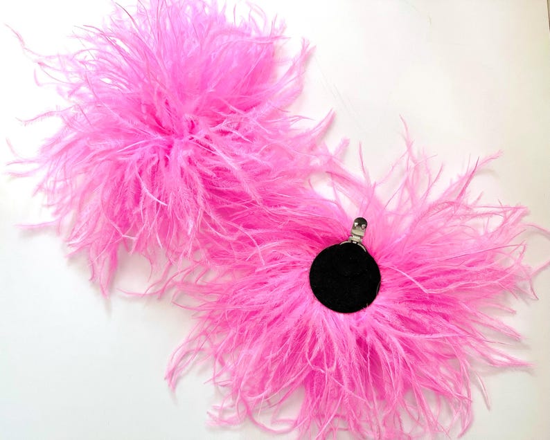 May include: Two bright pink feather accessories, each featuring a circular black base with a silver metal clip attached to the center. The soft, fluffy plumes radiate outward from the dark circular centers, creating a voluminous, textured appearance.