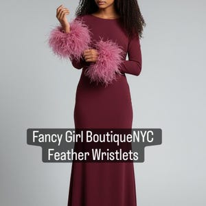 May include: A burgundy long-sleeve dress with a floor-length skirt. The model is wearing pink feather wristlets. The text on the image reads "Fancy Girl Boutique NYC Feather Wristlets."
