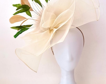 May include: A cream-colored fascinator with a large, layered flower design. The headpiece features a mesh overlay and is adorned with green, tan, and cream-colored feathers and leaves. The hat is mounted on a thin, brown headband.