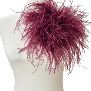 May include: A deep burgundy ostrich feather accessory featuring long, wispy plumes arranged in a dense, voluminous cluster. The feathers radiate outward from a central point, creating a textured, layered appearance with a rich, dark red color.