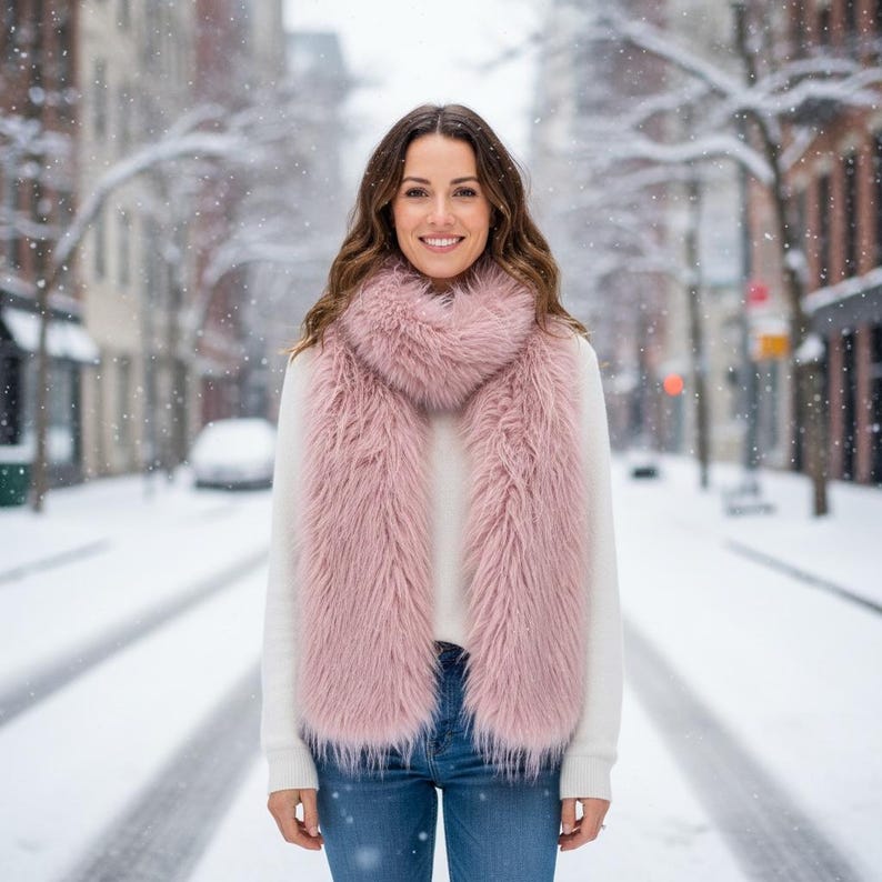 May include: A woman wearing a fluffy, light pink faux fur scarf over a white sweater and blue jeans. The scarf is long and rectangular, with a soft, textured appearance. The background shows a snowy street scene.