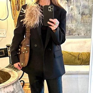 May include: A black double-breasted blazer featuring a large, beige feather embellishment on the left lapel. The blazer is paired with dark trousers and a brown leather shoulder bag with gold-tone metal studs and a wide strap.