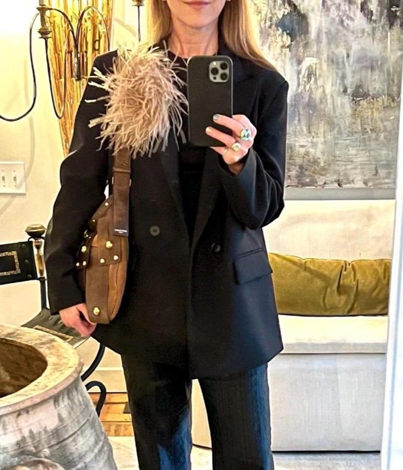 May include: A black double-breasted blazer featuring a large, beige feather embellishment on the left lapel. The blazer is paired with dark trousers and a brown leather shoulder bag with gold-tone metal studs and a wide strap.