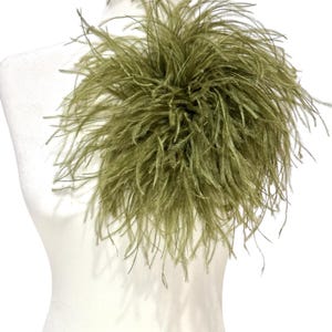 May include: A decorative accessory featuring a dense cluster of olive green ostrich feathers. The long, wispy plumes are arranged in a circular, voluminous shape, creating a textured and layered appearance that radiates outwards from the centre point.