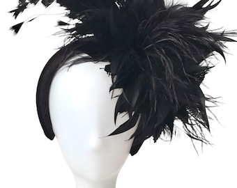 May include: A black feather headband, displayed on a white mannequin head. The headband features a thick, black band and a large, elaborate arrangement of black feathers. The feathers are layered and create a voluminous, textured effect, suitable for special occasions.
