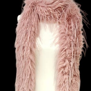 May include: A dusty rose faux fur scarf displayed on a white mannequin. The scarf is long and fluffy, with a soft, textured appearance. The faux fur has a slightly uneven texture, adding to its visual appeal. The background is black.