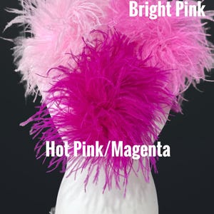 May include: Three fluffy ostrich feather clusters displayed on a white mannequin head. The feathers are labeled Pale Powder Pink, Bright Pink, and Hot Pink/Magenta. The soft, wispy plumes are arranged in distinct shades of pink, creating a textured look.