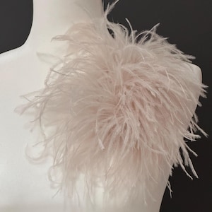 May include: A pale blush pink ostrich feather accessory attached to a white mannequin shoulder. The piece features long, wispy, and delicate plumes that create a voluminous, textured appearance with a soft, light-reflecting quality throughout.