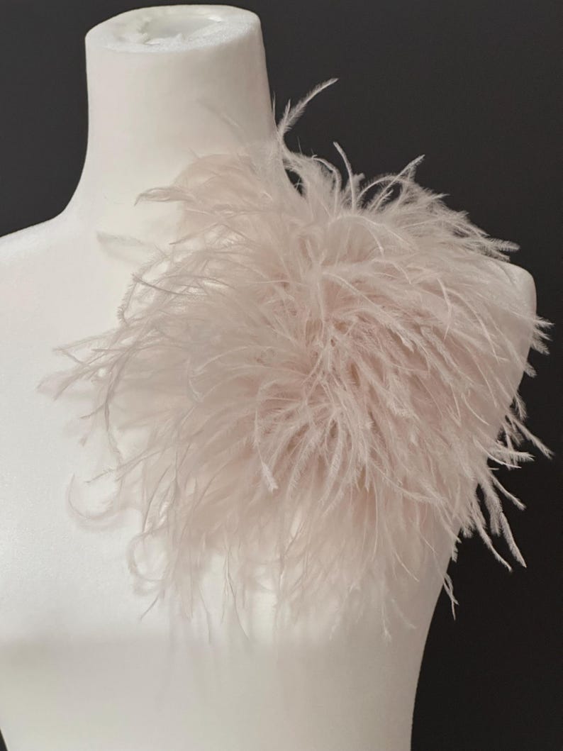 May include: A pale blush pink ostrich feather accessory attached to a white mannequin shoulder. The piece features long, wispy, and delicate plumes that create a voluminous, textured appearance with a soft, light-reflecting quality throughout.