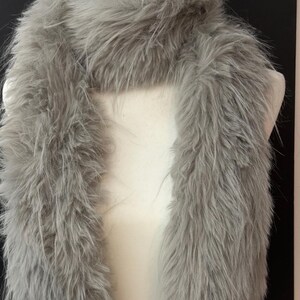 May include: A fluffy, gray faux fur scarf is draped over a white mannequin. The scarf has a long, rectangular shape and is made of soft, textured material. The scarf is a light gray color and appears to be warm and cozy.