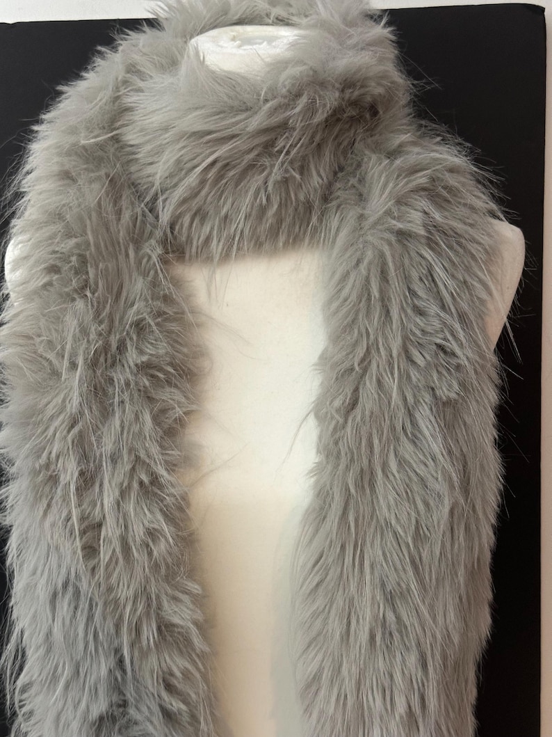 May include: A fluffy, gray faux fur scarf is draped over a white mannequin. The scarf has a long, rectangular shape and is made of soft, textured material. The scarf is a light gray color and appears to be warm and cozy.